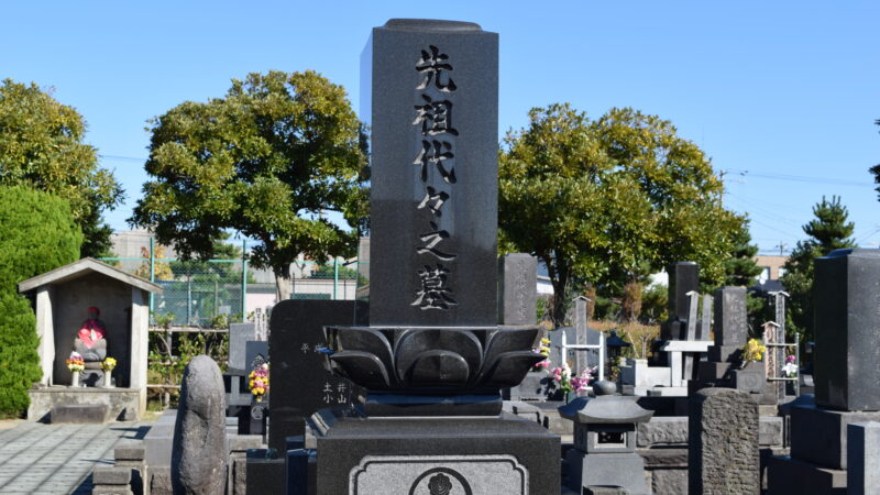 晴れた空の下に建つ「先祖代々之墓」と刻まれた黒い和型墓石と周囲の墓地の風景
